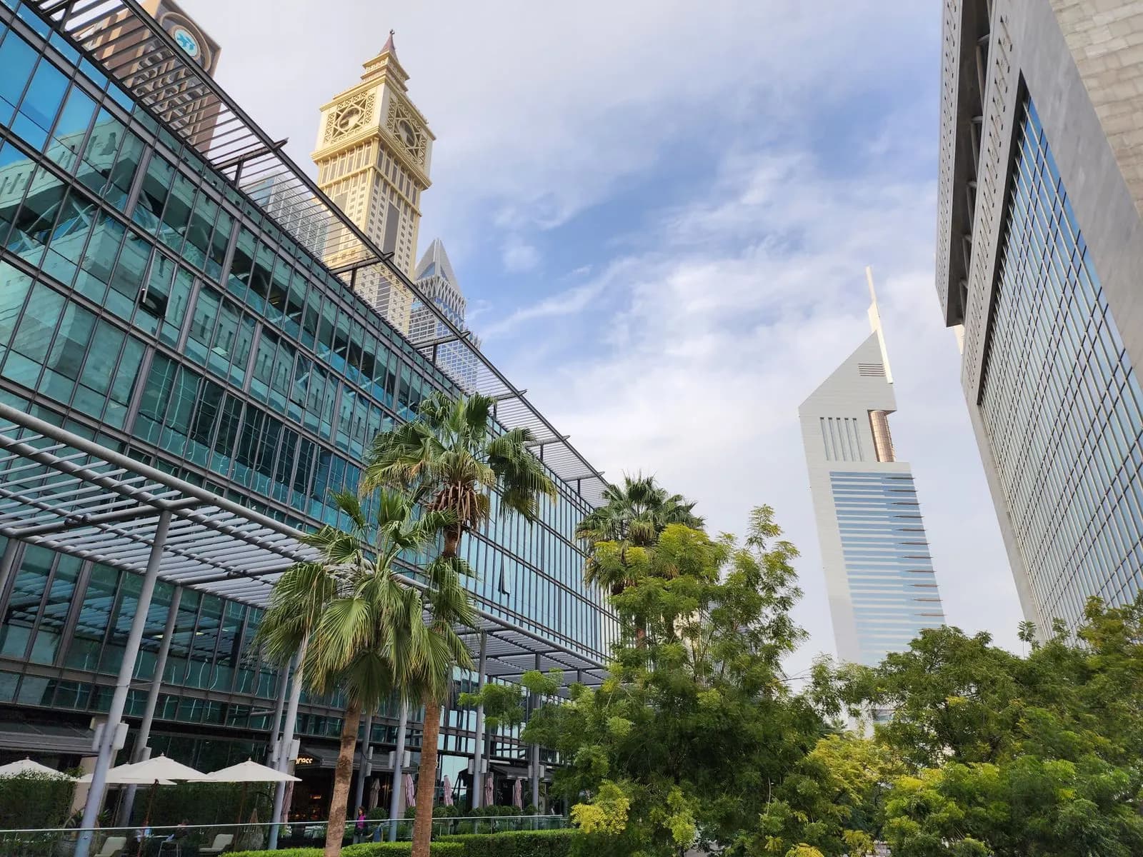 أبراج الإمارات ترتفع خلف مبانٍ مكتبية زجاجية منخفضة وأشجار نخيل.