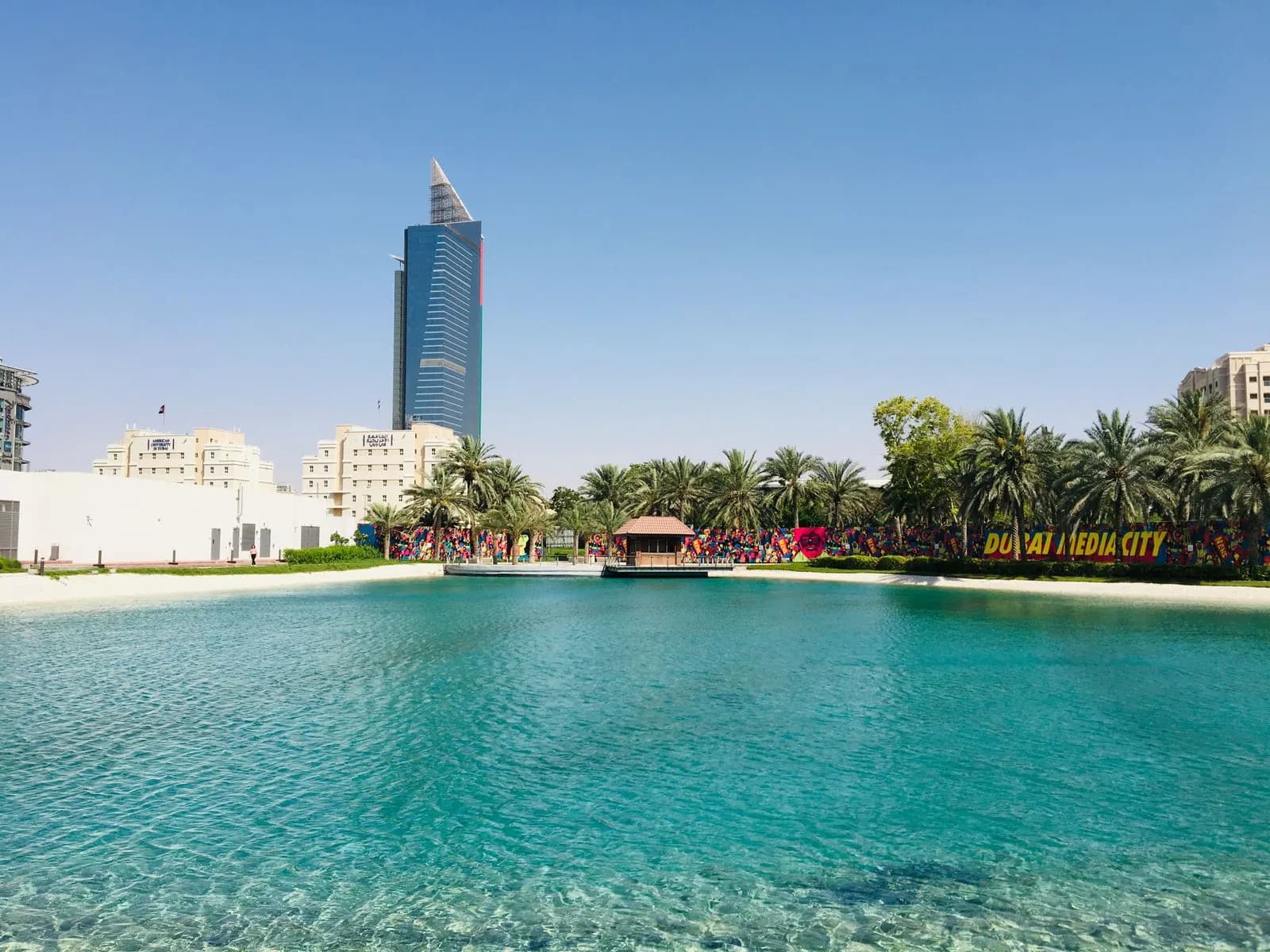 عبارة DUBAI MEDIA CITY على جدار خلف بحيرة فيروزية وأشجار نخيل، ويرتفع خلفها برج أزرق.